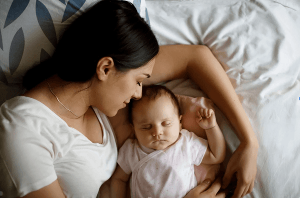 Mother lying in bed peacefully holding her sleeping baby, both resting on white bedding
