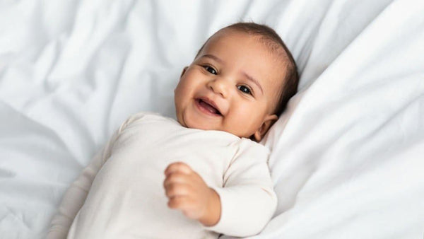 Smiling baby in white onesie lying on soft white bedding, looking happy and healthy