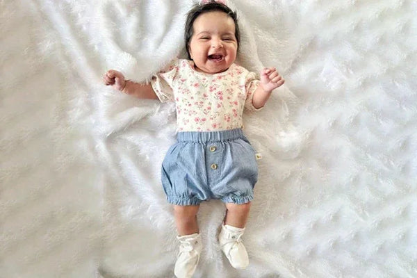 Smiling baby girl lying on a white fluffy blanket wearing floral top, blue shorts, and white booties