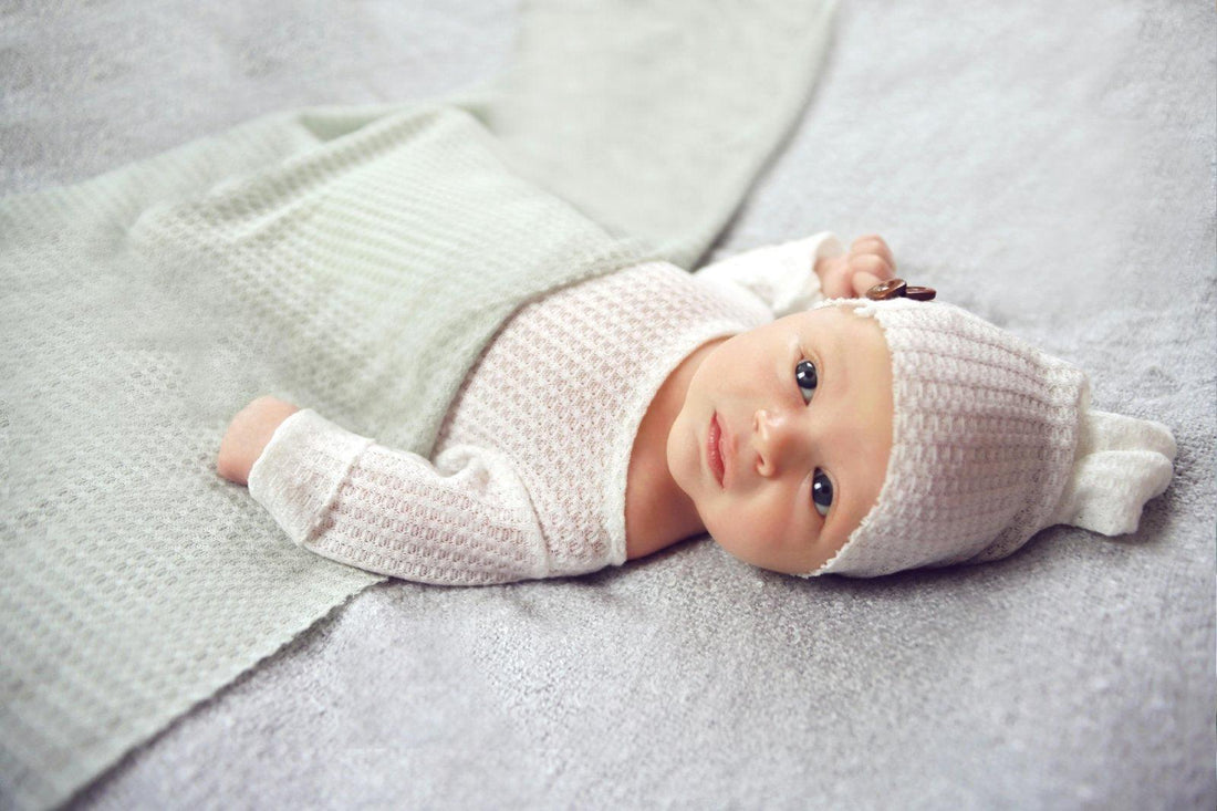 Newborn baby lying on gray blanket wearing knitted hat and white cozy outfit