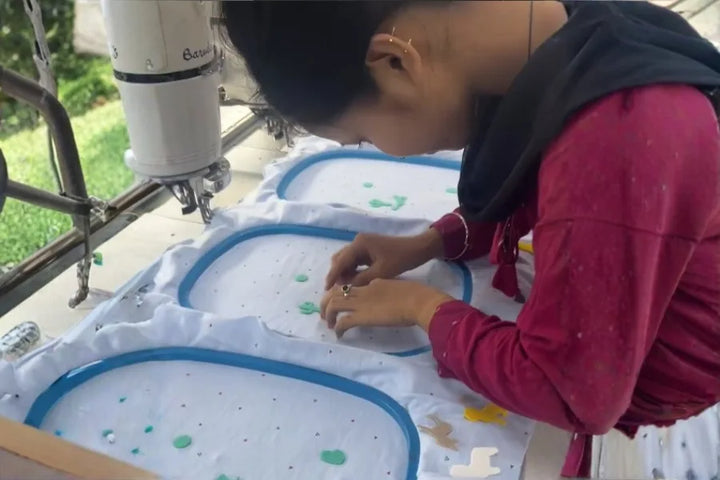 Woman in maroon shirt hand embroidering fabric stretched in blue hoops on a sewing machine table