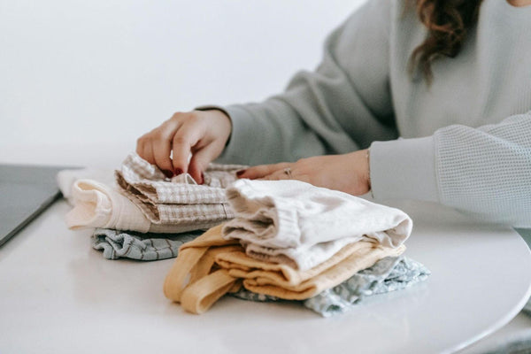 Person folding soft, neutral-colored baby clothes on white table in cozy indoor setting