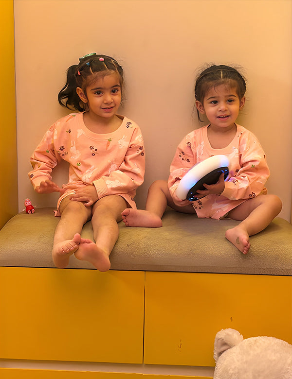 Two young girls sitting on a cushioned bench in peach pajama sets, one holding a light-up toy, with yellow drawers below