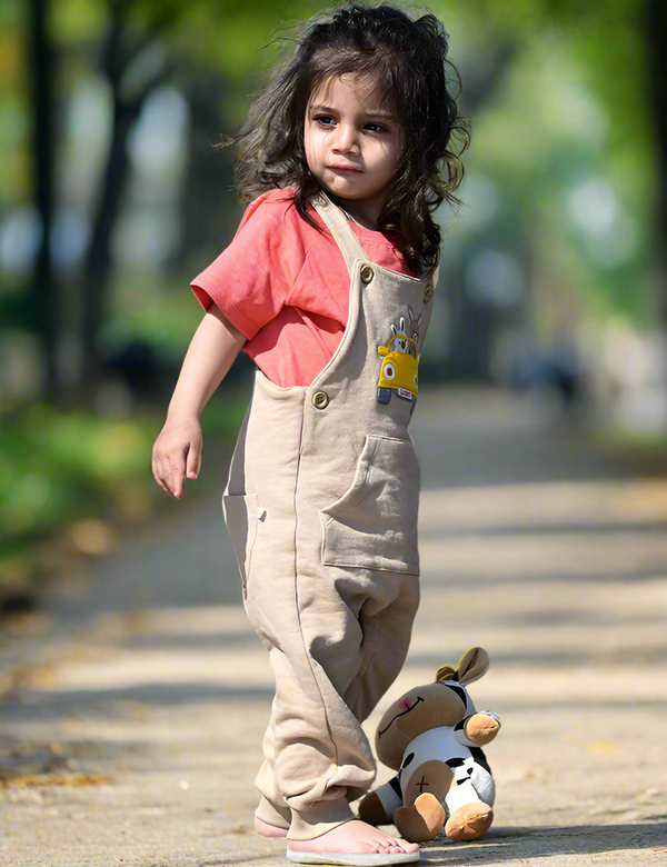 A child wearing a red tee and beige overalls standing next to a stuffed toy on a couch.