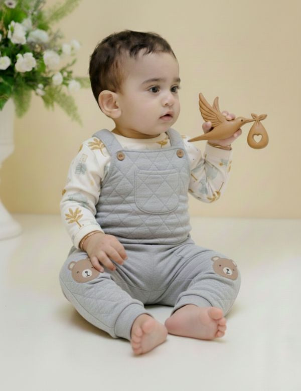 Baby wearing grey quilted overalls with bear patches, holding wooden bird toy, sitting indoors