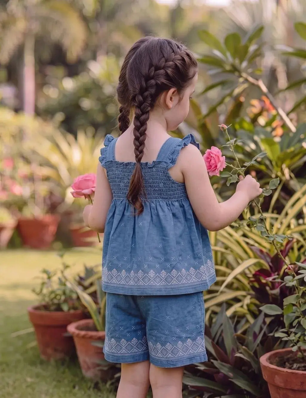 Back view of a young girl model with braided hair in a garden holding pink roses, wearing an organic cotton denim blue smocked top and shorts set with white embroidery.