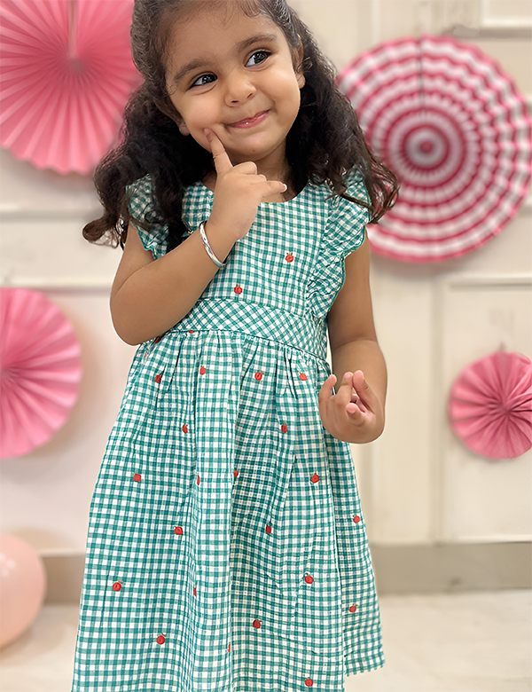 Little girl wearing a green gingham dress with apple embroidery and ruffle sleeves.