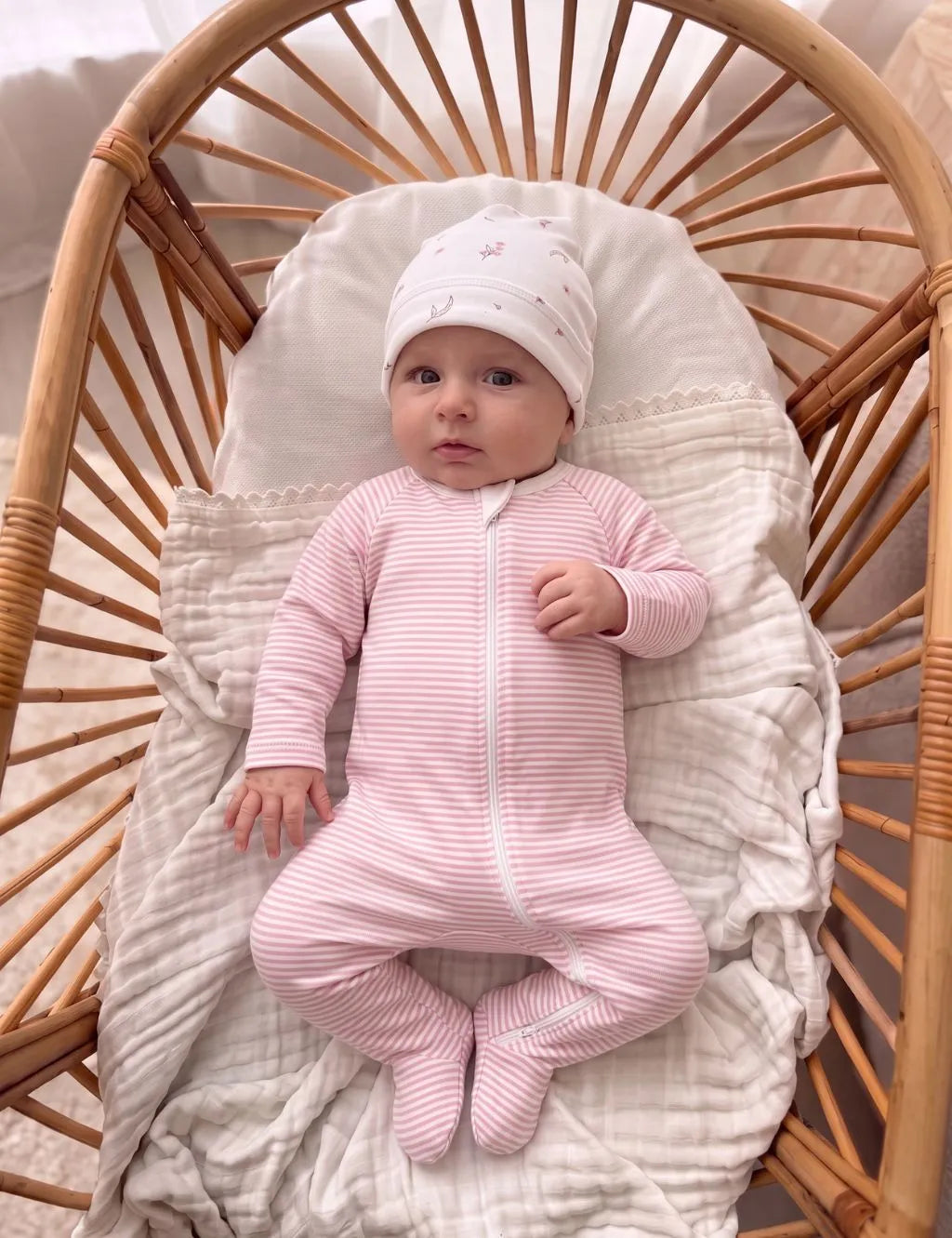 Baby wearing a pink striped organic cotton zip romper and matching hat in a rattan bassinet