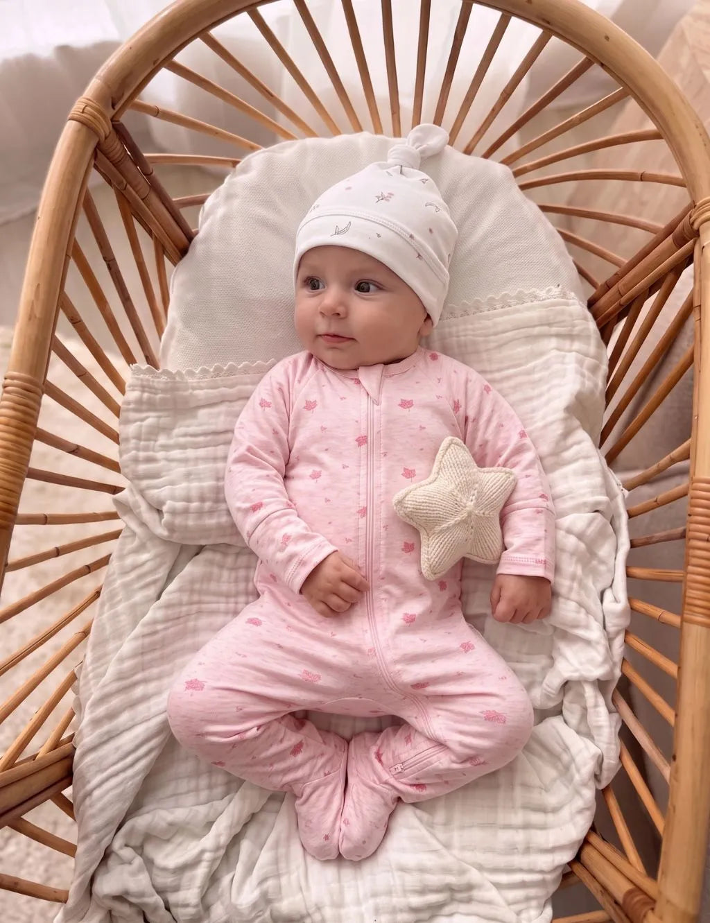 Baby wearing a pink organic cotton zip romper and white hat in a rattan bassinet