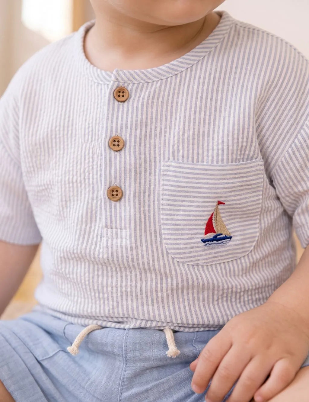 A close-up of a child wearing a short-sleeve white Henley shirt with thin gray vertical stripes, three brown buttons, and a chest pocket featuring a small embroidered sailboat motif. The child is also wearing light blue shorts.