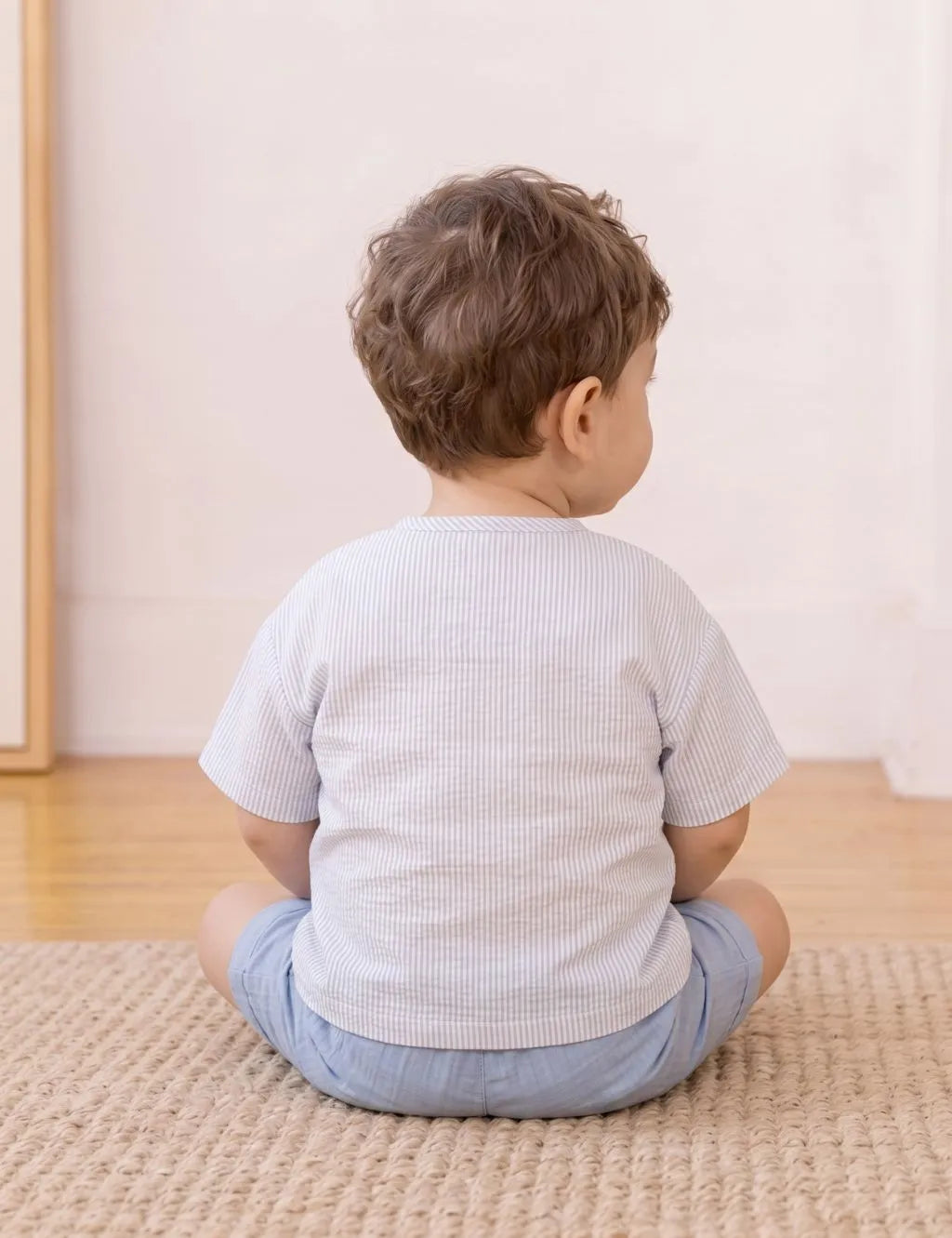 A toddler boy sitting on a woven rug, viewed from behind, wearing a white waffle-knit short-sleeve shirt and light blue shorts in a bright, minimalist room.