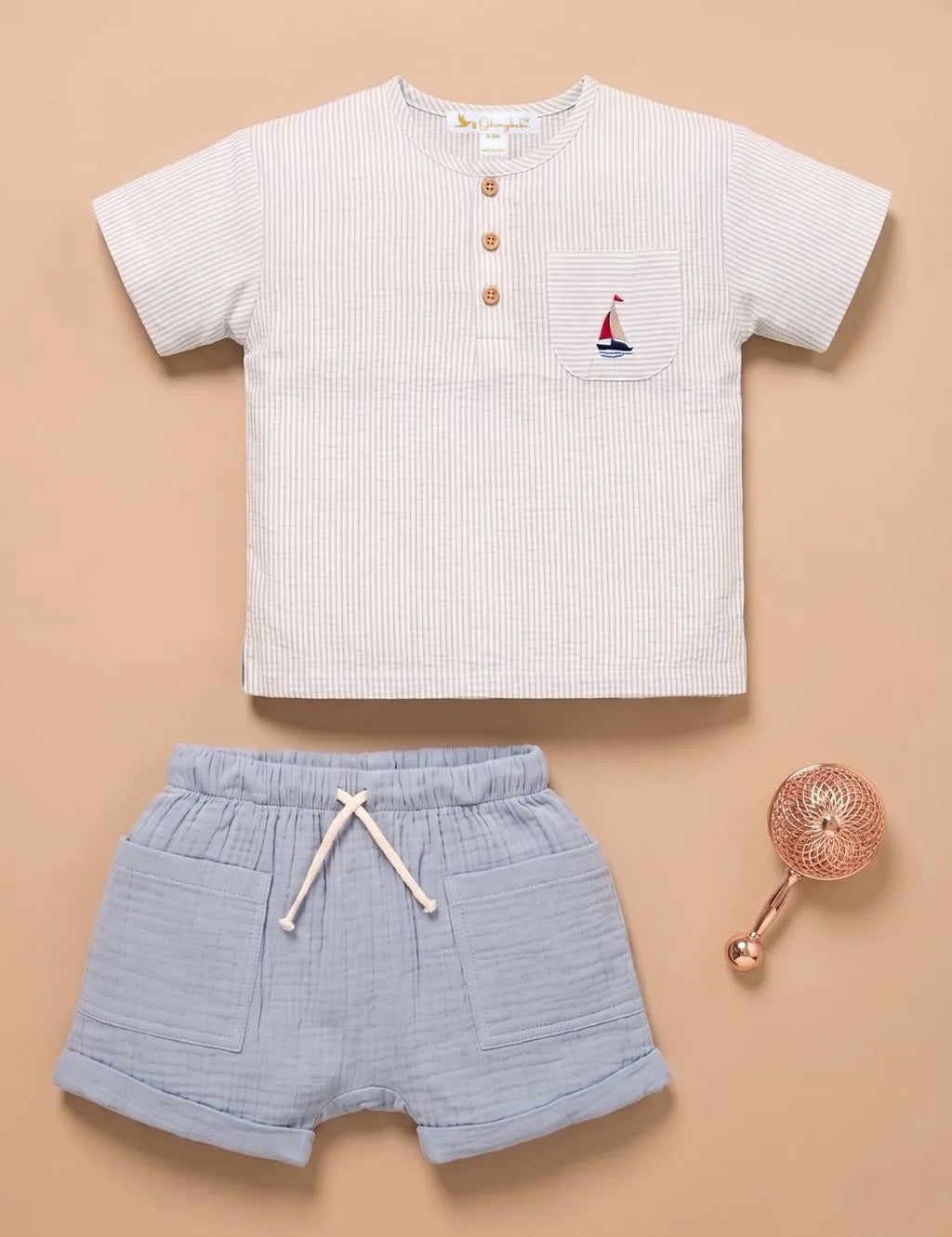 A baby boy's two-piece outfit set consisting of a short-sleeve henley shirt in white and navy micro-stripe fabric with a small sailboat chest pocket, paired with light blue textured shorts with a white drawstring, displayed on a beige background with a small rose gold baby rattle prop.