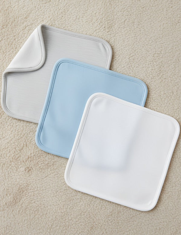 Three square microfiber cleaning cloths in white, blue, and gray on a beige textured surface