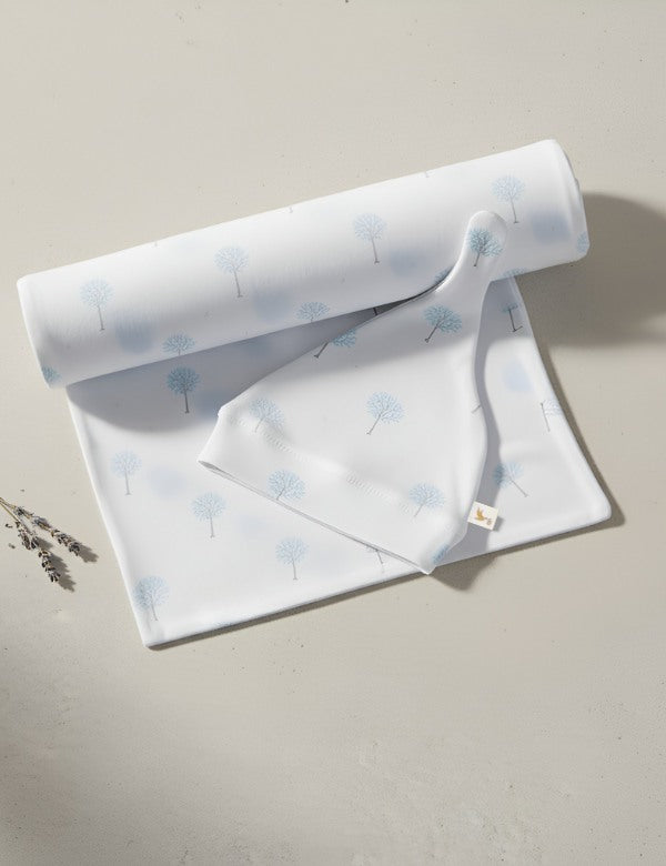 White baby blanket with blue tree prints rolled and folded on beige surface with dried flowers