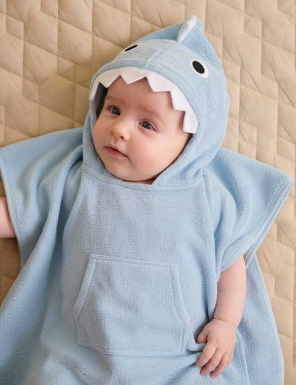 baby wearing a light blue shark-themed hooded towel lying on a quilted beige surface