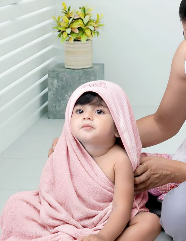 Cute baby wrapped in pink towel with hood, sitting indoors near white railing and potted plant