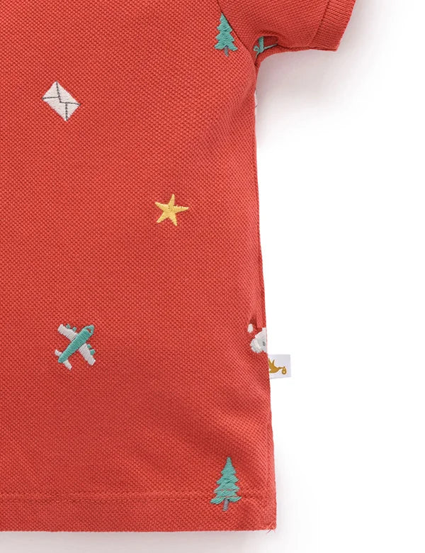 Red polo shirt with airplane, star, and tree patterns on a white background
