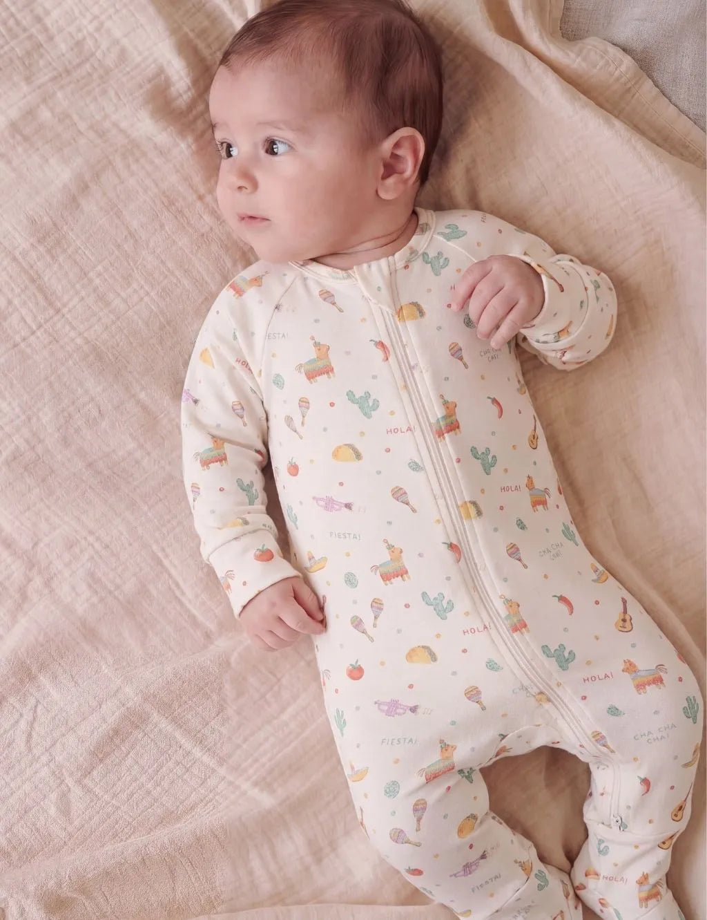 Baby lying down wearing a cream zip-up romper featured in a fiesta print with piñatas, cacti, and tacos