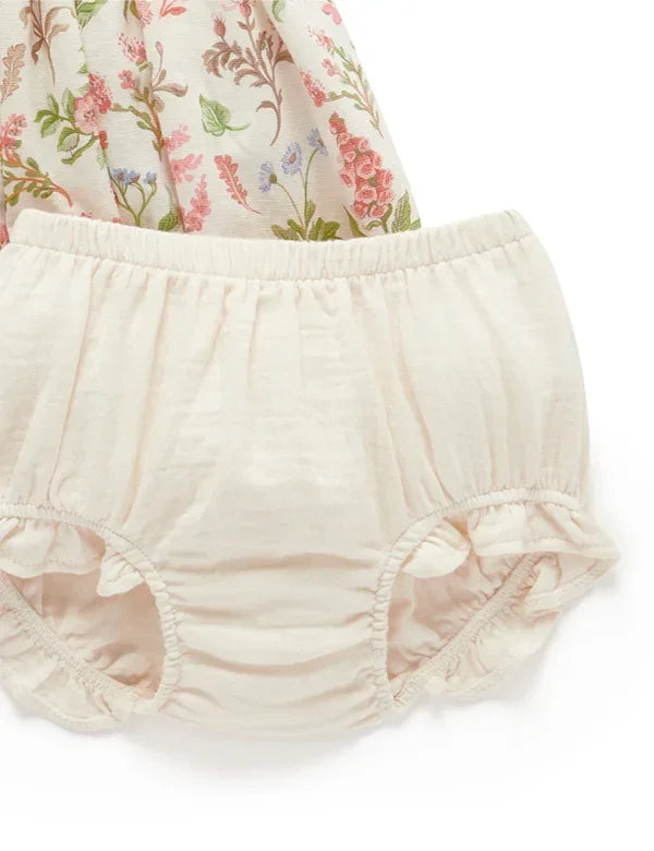 Cream-colored baby bloomer shorts with elastic waist and ruffled leg openings, floral fabric background