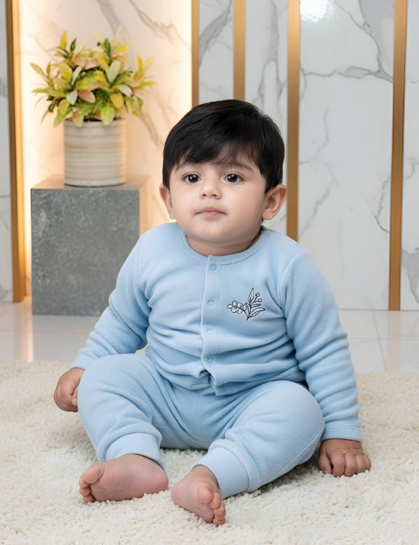 Cute baby boy in blue onesie sitting on white rug with potted plant and marble wall background