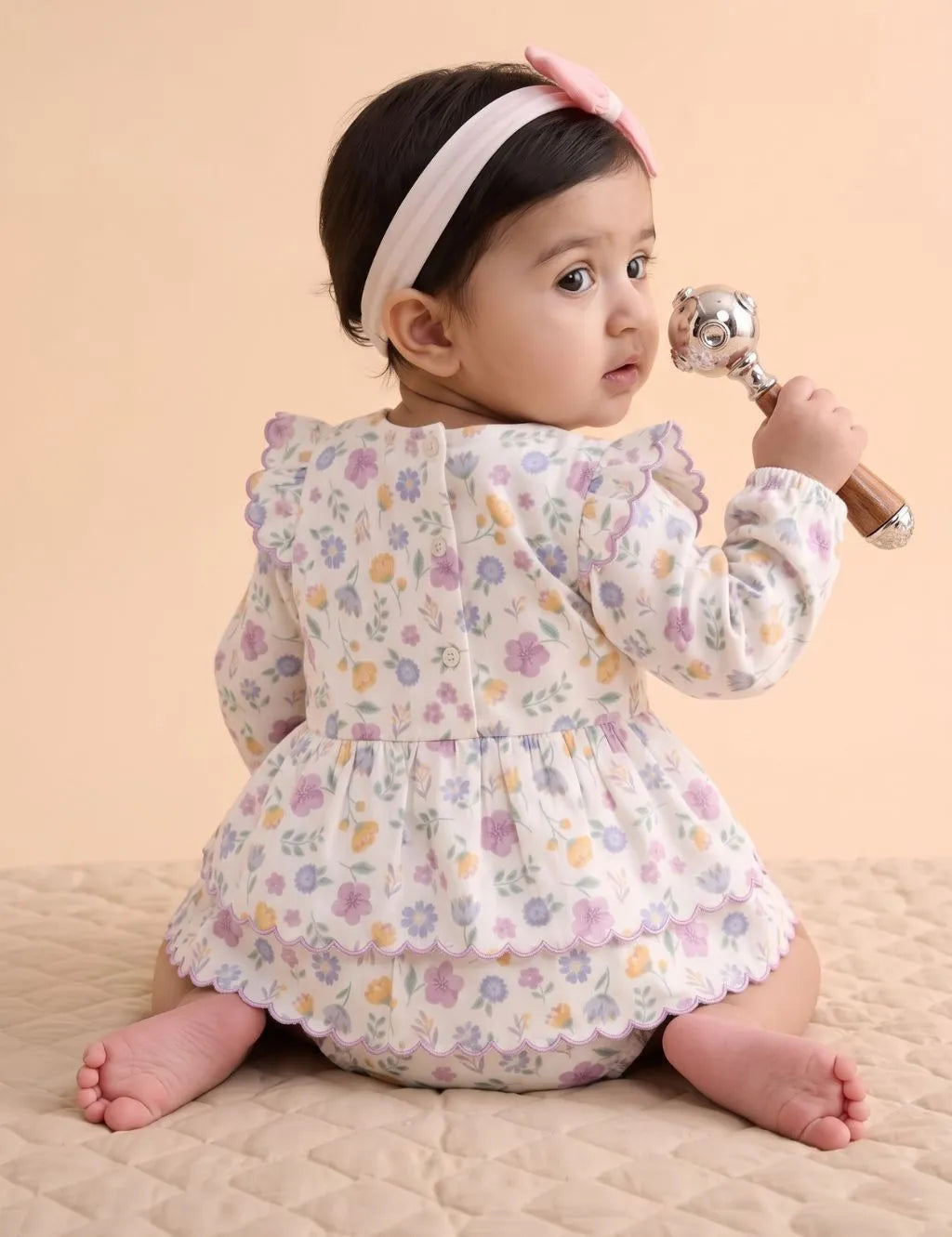 Back view of a baby girl sitting with a rattle wearing a scalloped floral tiered dress romper with back button closures.