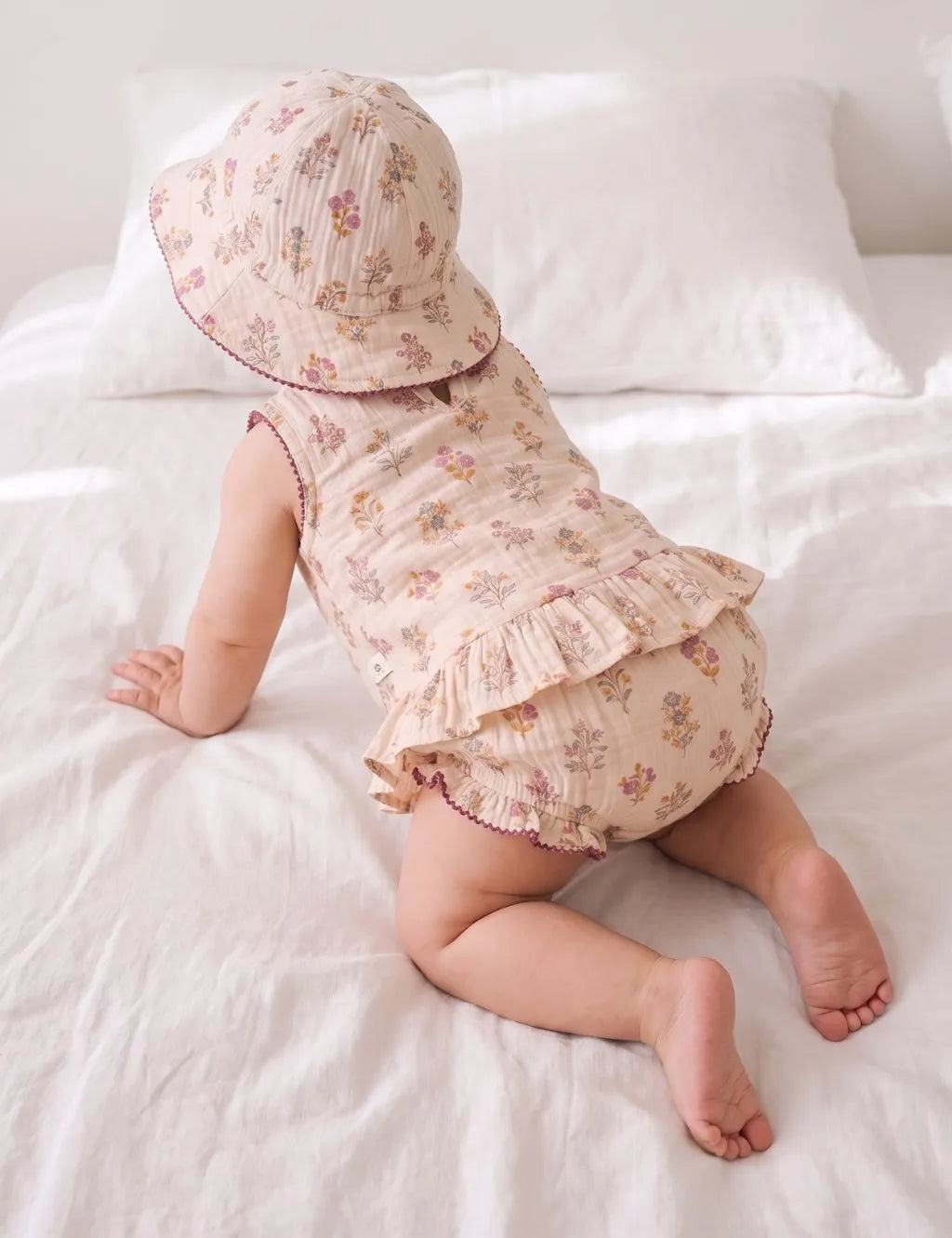 Back view of a crawling baby girl wearing a floral ruffle top, matching bloomers, and a sun hat on white bedding.