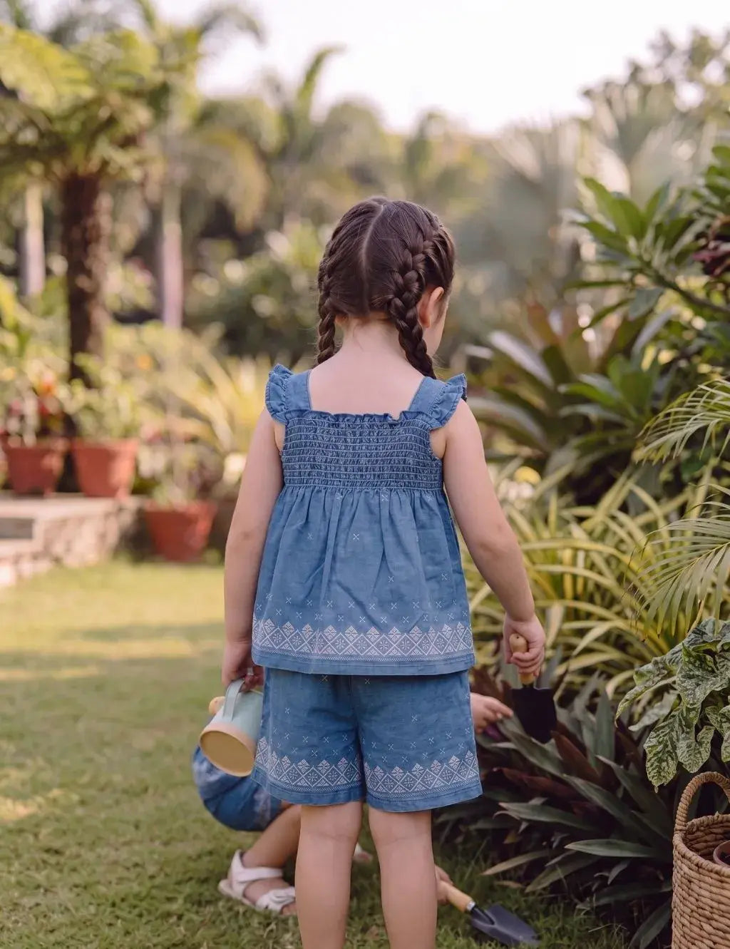 Back view of a young girl model with braided hair gardening outdoors, wearing an organic cotton denim blue smocked top and shorts set with white embroidery.