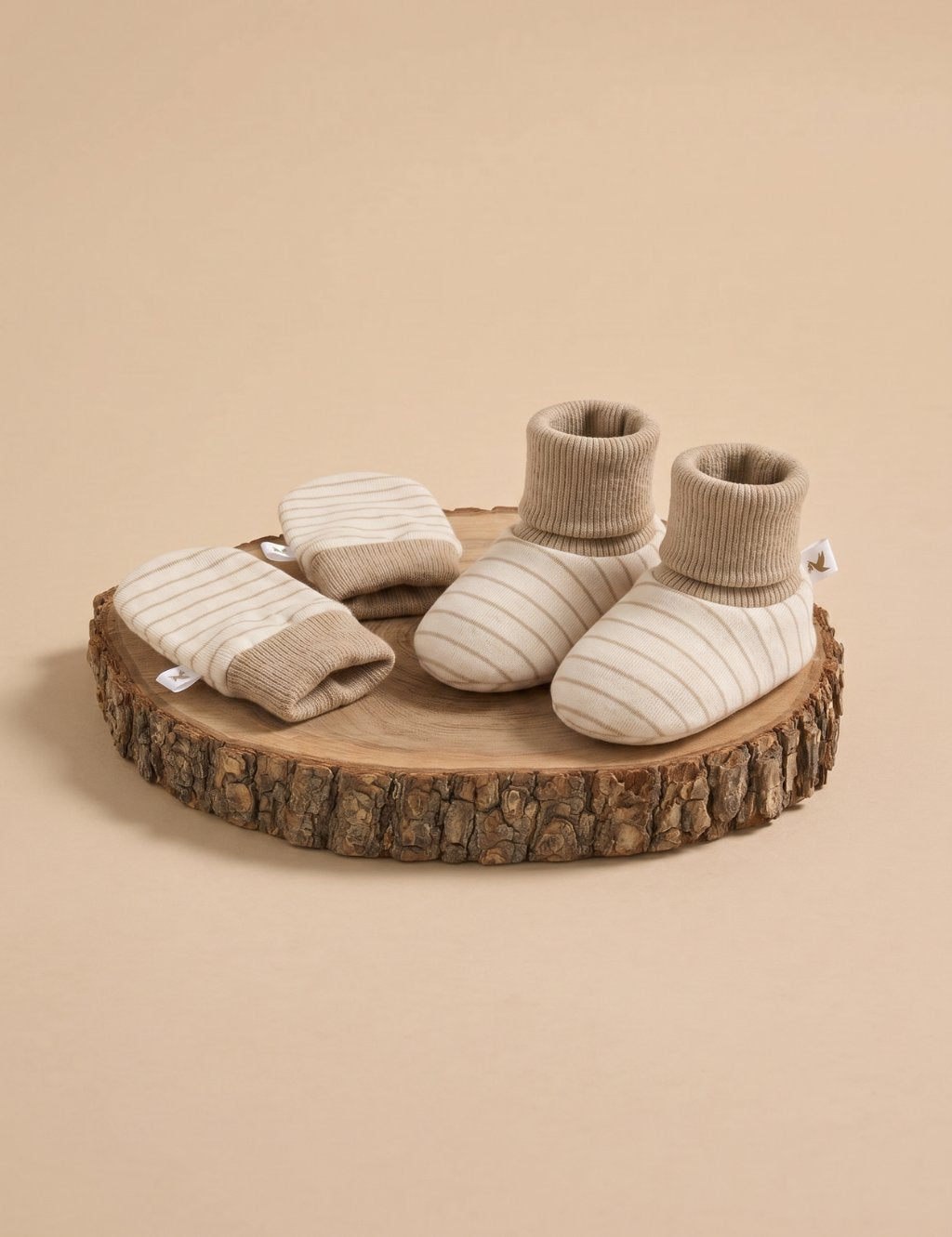 A matching set of cream-colored baby booties and scratch mittens featuring thin beige stripes and solid ribbed cuffs, displayed on a rustic wooden log slice against a plain beige background.