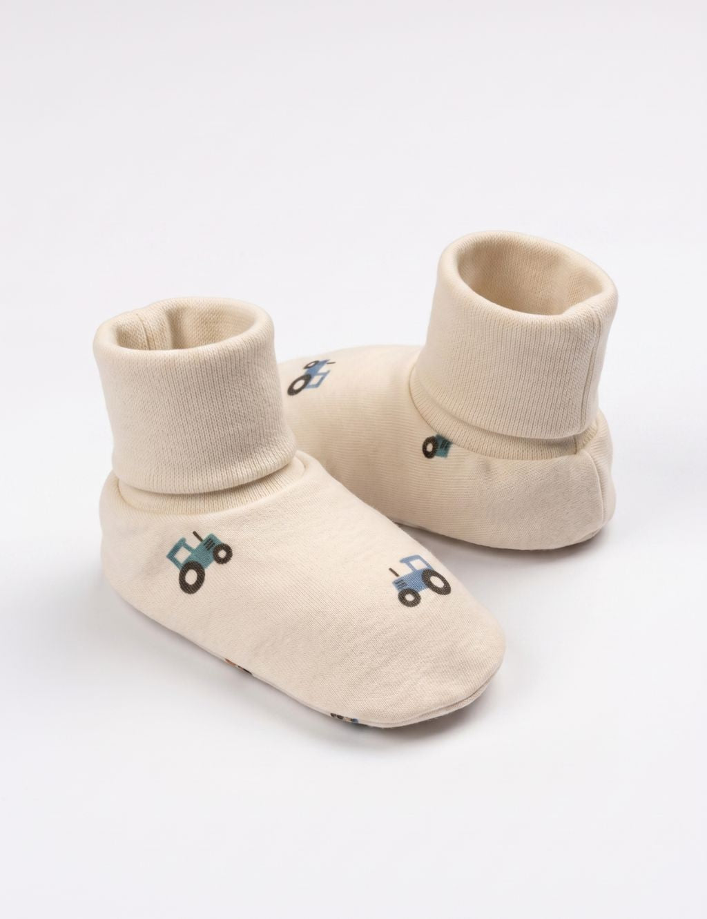 A pair of soft cream-colored baby booties featuring a small blue tractor print, displayed against a plain white background.