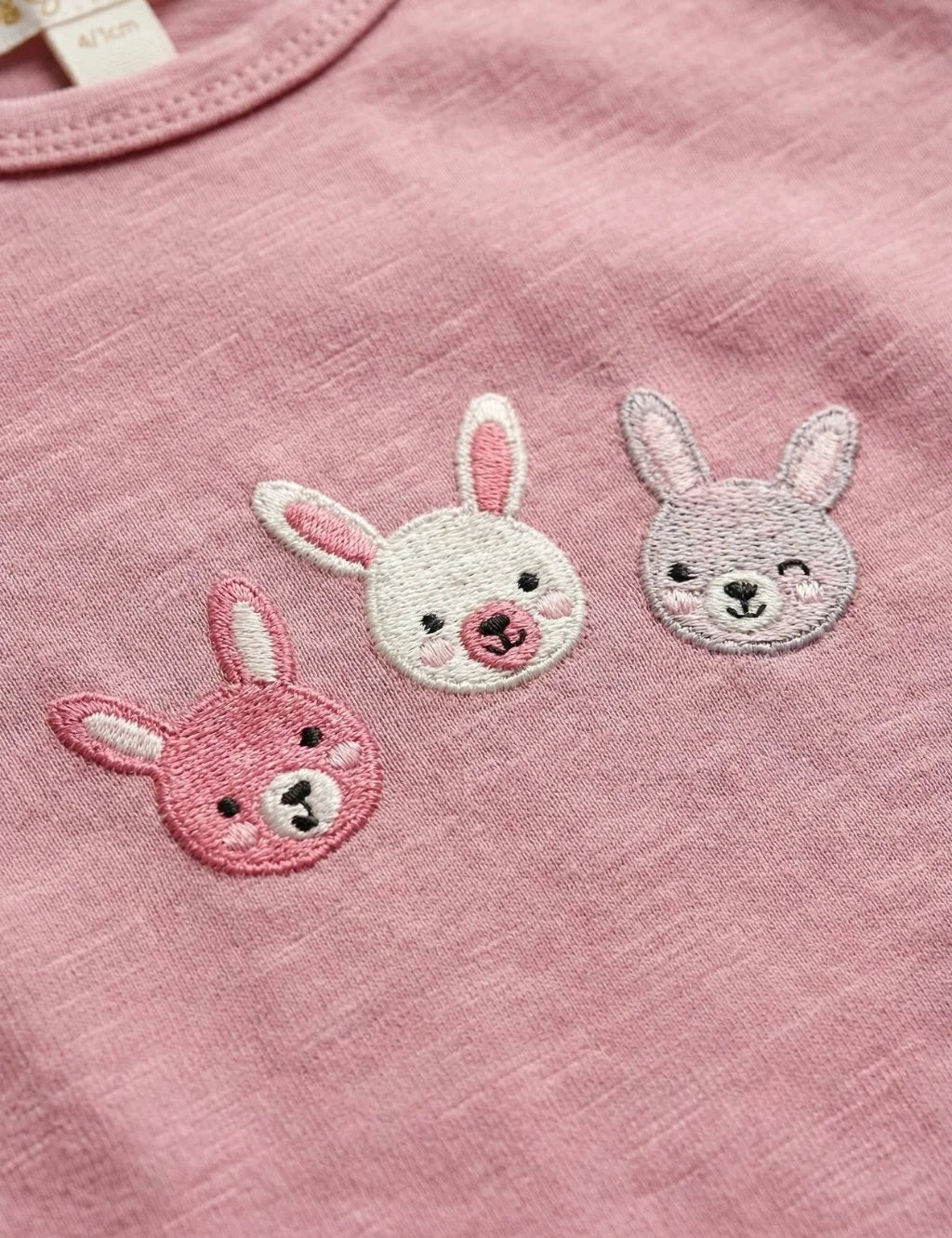 Close-up detail of three embroidered bunny faces in shades of pink on an organic cotton baby tee.