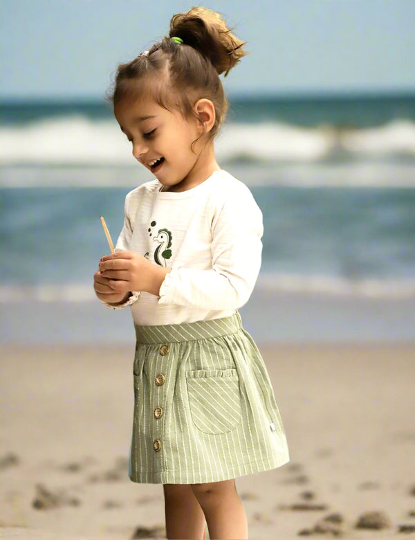 A young child, who appears to be of South Asian descent, on a beach holding a stick, wearing a butterfly applique long-sleeve tee and striped safari skirt. Shop adorable kids' sets India.