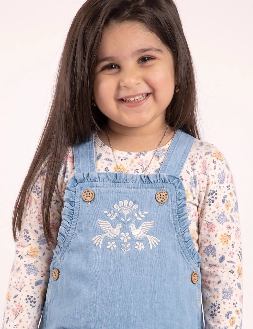 Close-up of a smiling toddler girl in a denim short romper featuring bird embroidery and ruffle bib detail.