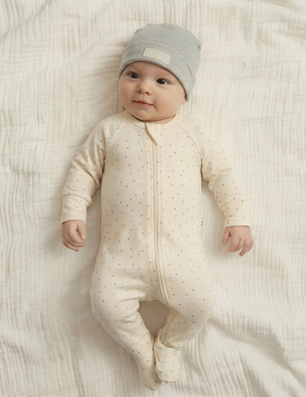 Baby in a cream-colored onesie with subtle pattern and matching hat, lying on a soft textured surface.