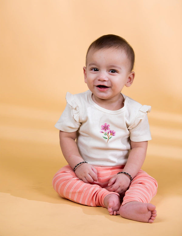A joyful baby, who appears to be of Caucasian descent, sitting and smiling while wearing a white bodysuit with floral embroidery and striped leggings. Shop adorable baby clothes India.
