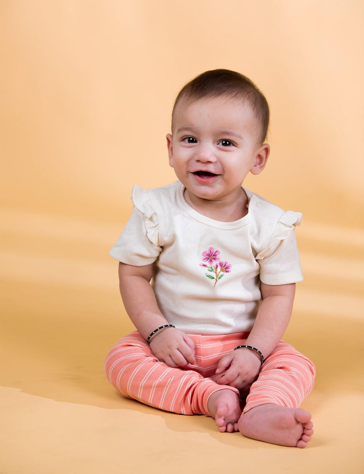 A joyful baby, who appears to be of Caucasian descent, sitting and smiling while wearing a white bodysuit with floral embroidery and striped leggings. Shop adorable baby clothes India.