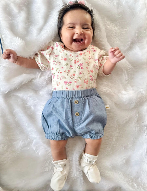 A happy baby, who appears to be of South Asian descent, lying on a white blanket wearing a floral print top and blue denim bloomers. Shop adorable baby sets India.