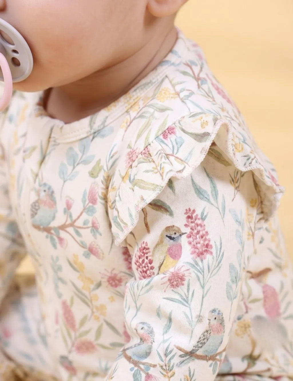 Close-up of a baby's botanical bird print romper detailing the shoulder ruffle and soft cotton fabric