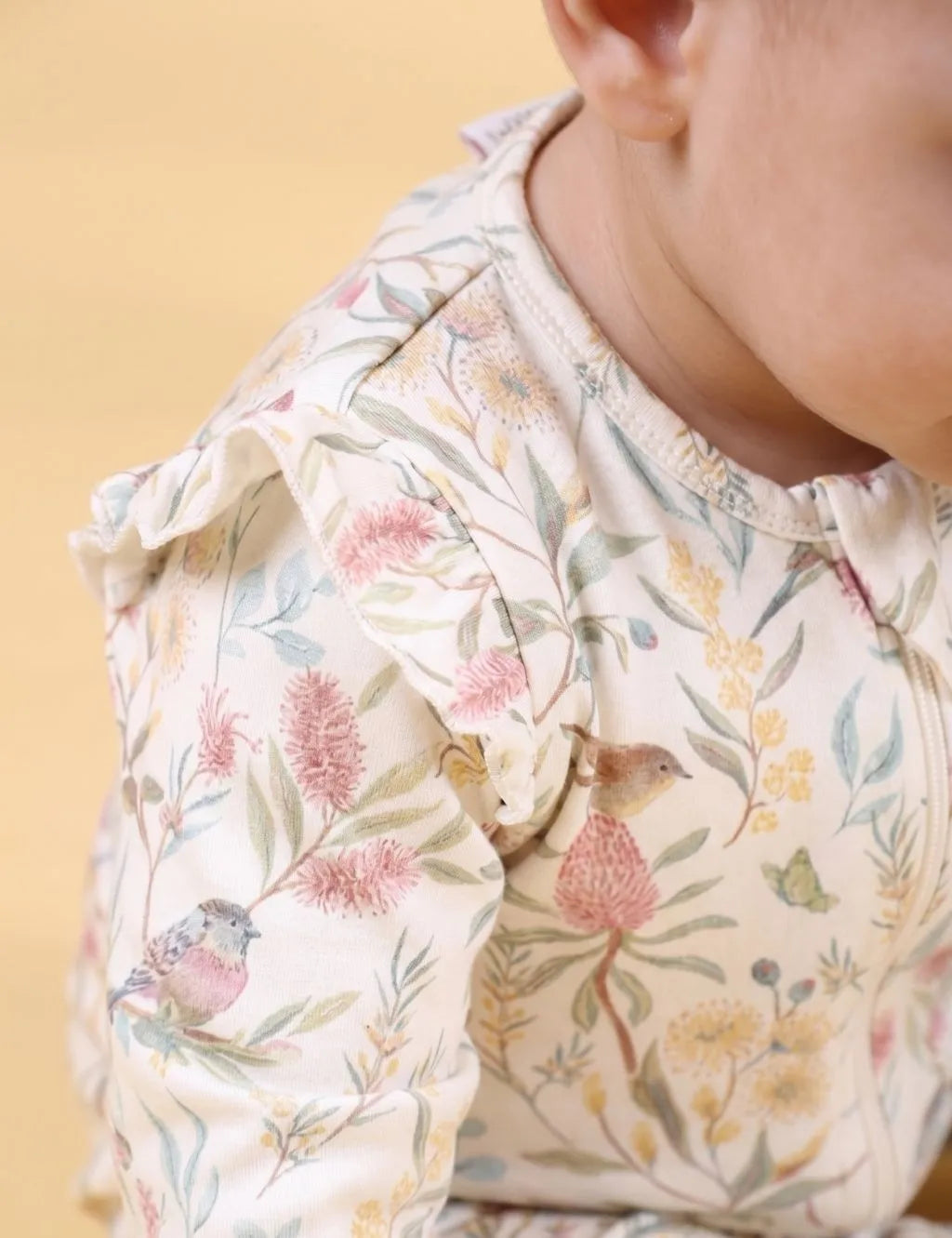 Close-up side view of a baby wearing a botanical floral and bird print romper showing the ruffle shoulder sleeve.
