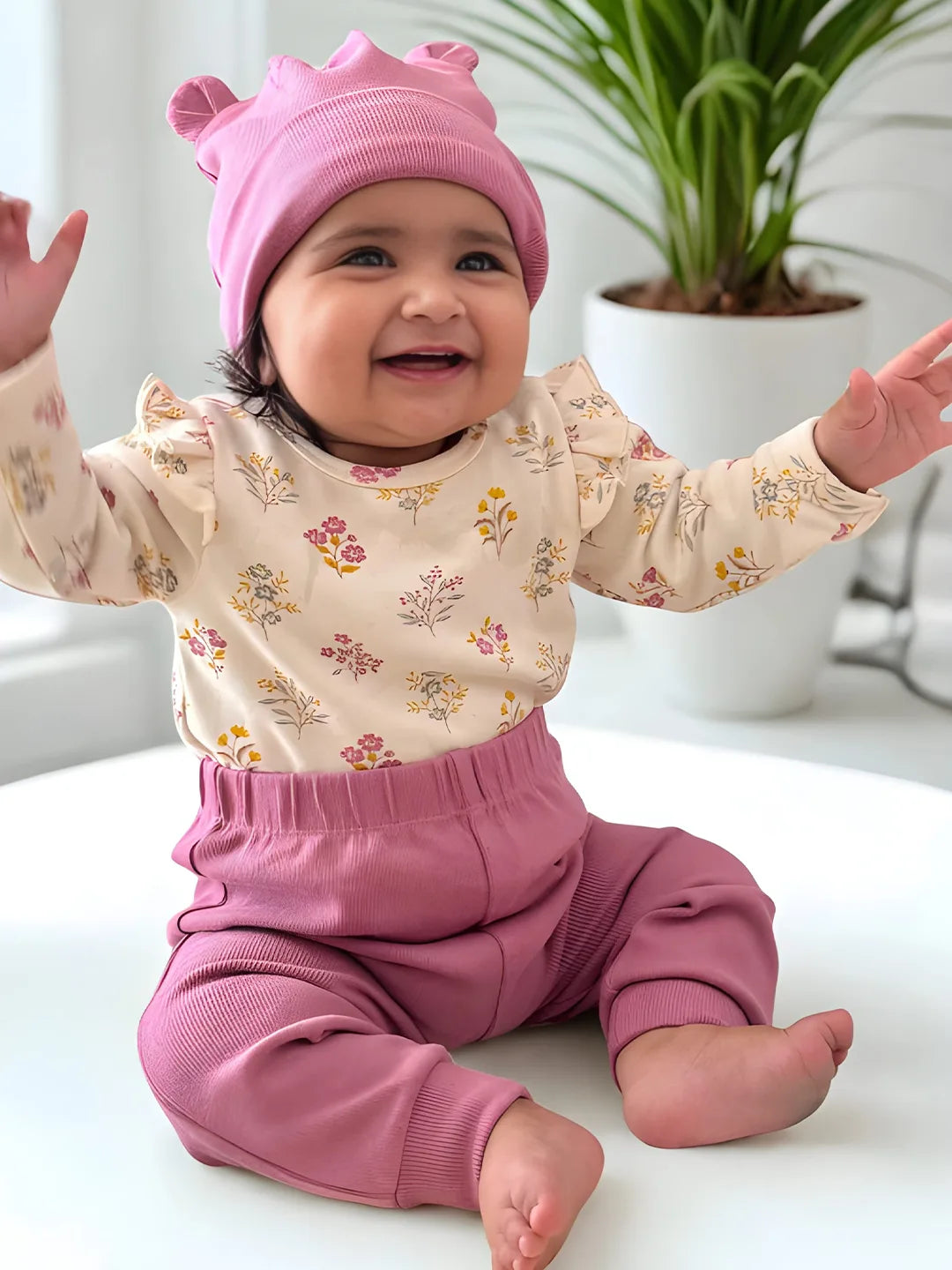 Happy baby in floral cream top, pink pants, and pink hat sitting indoors with green plant