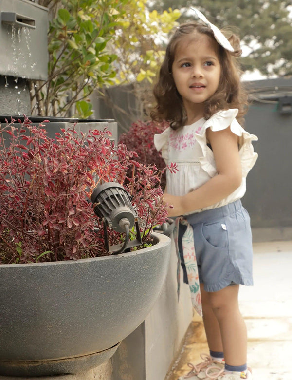 A young girl, who appears to be of South Asian descent, standing near a water feature, wearing an embroidered white tunic top and denim-look shorts with a floral sash. Shop cute baby sets India.