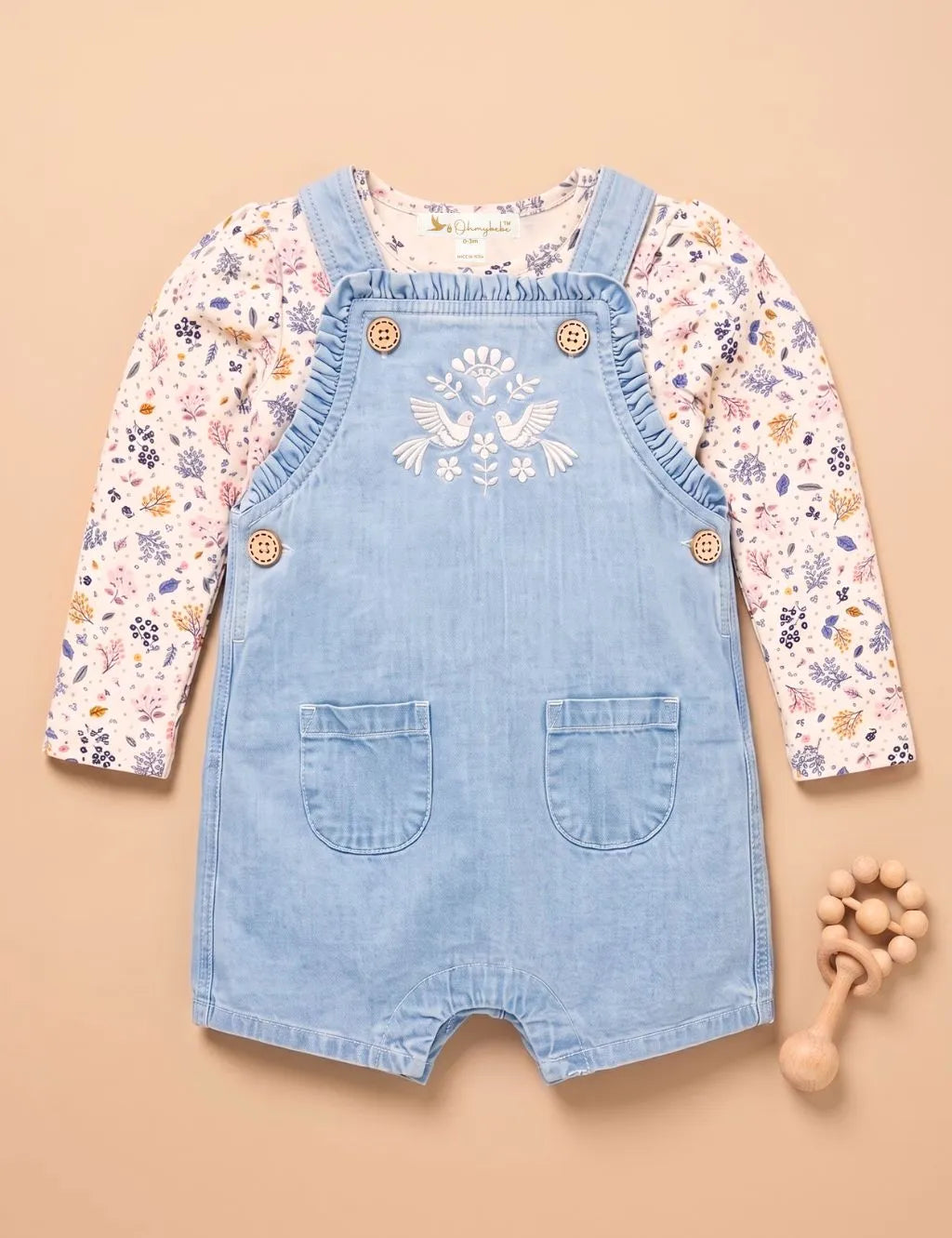 Flatlay of a light wash denim embroidered short romper paired with a floral long-sleeve top and wooden teether.