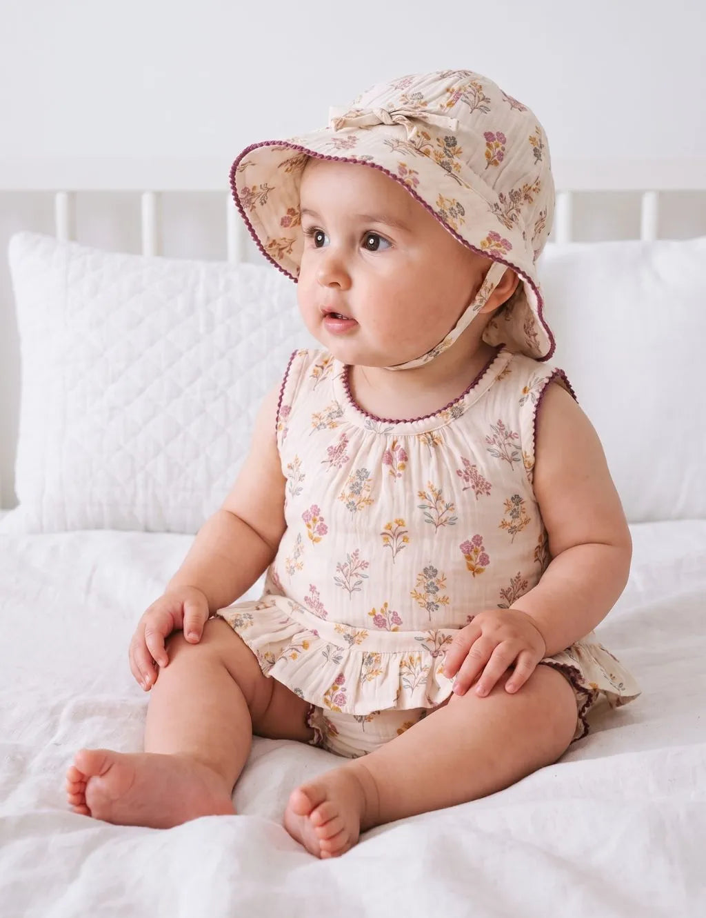 Baby girl sitting on a bed wearing a floral sleeveless ruffle romper and matching wide-brim sun hat.