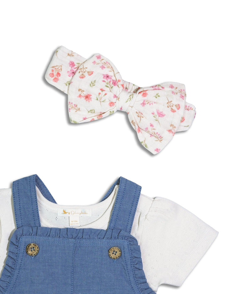 Close-up showing the white headband with a pink floral bow and the denim dungaree with wooden buttons and a matching floral bow detail. See cute baby accessories India.