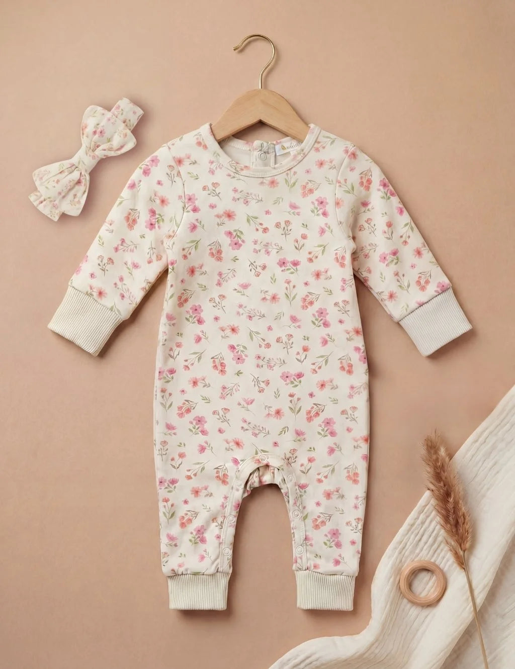 Full front view of a white organic cotton long-sleeve footless baby growsuit featuring a delicate pink floral print, laid flat on a wooden hanger next to a matching bow headband