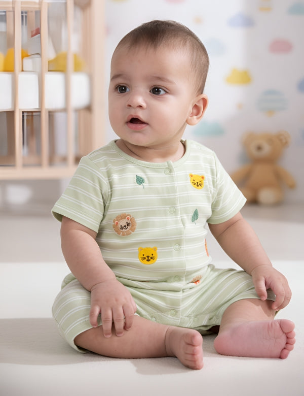 Baby boy in green striped romper with animal patches sitting on floor in nursery with crib and teddy bear