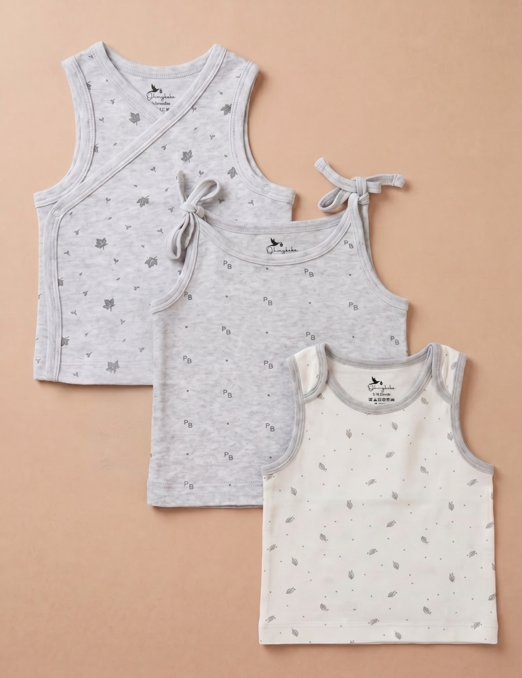 Flat lay view of three baby tank tops arranged diagonally in a set. From back to front: a light grey heather wrap-style tank top featuring a delicate scattered dark grey leaf pattern, a light grey heather camisole tank top patterned with small letters and dots finished with shoulder ties, and a cream-colored sleeveless tank top with a small grey leaf print and contrasting grey trim, all displayed against a plain beige background.