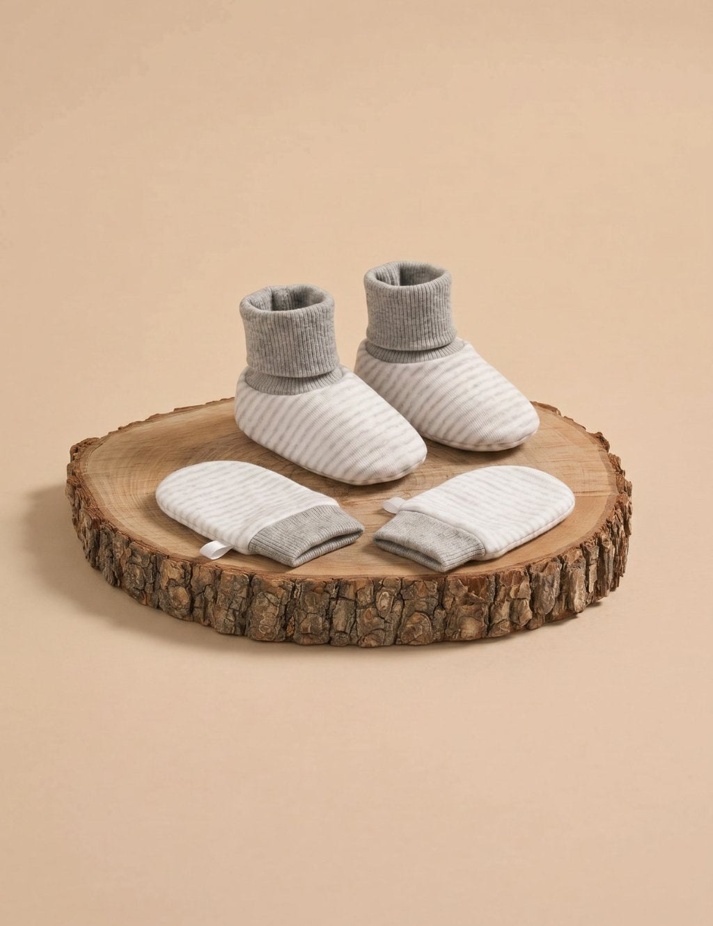 A matching set of cream-colored baby booties and scratch mittens featuring thin grey stripes and solid grey ribbed cuffs, displayed on a rustic wooden log slice against a plain beige background.