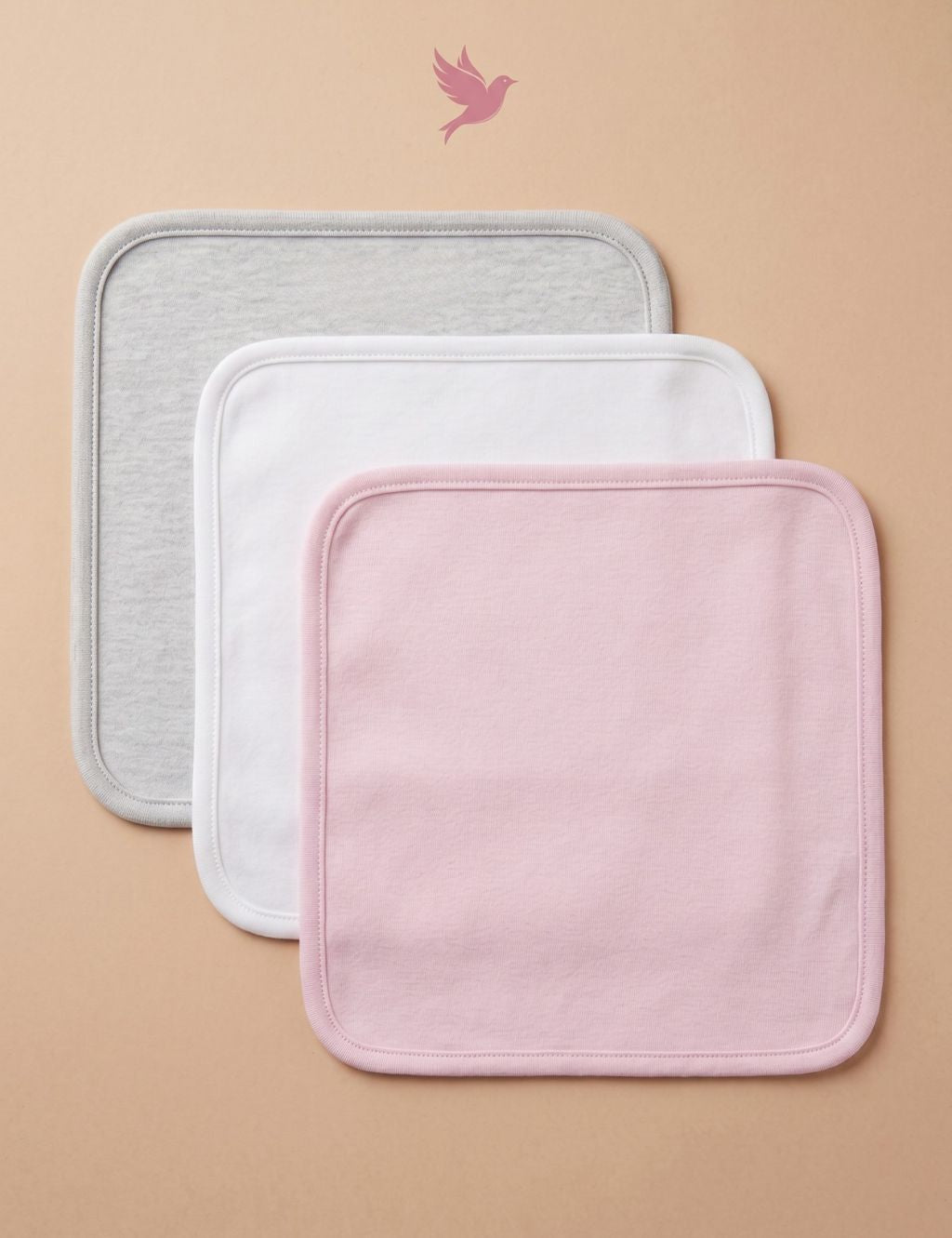 Flat lay view of three square baby washcloths arranged diagonally under a pink bird logo. From top to bottom: a solid light grey heather washcloth, a solid white washcloth, and a solid light pink washcloth, all featuring soft rounded corners and stitched edges, displayed against a plain beige background.