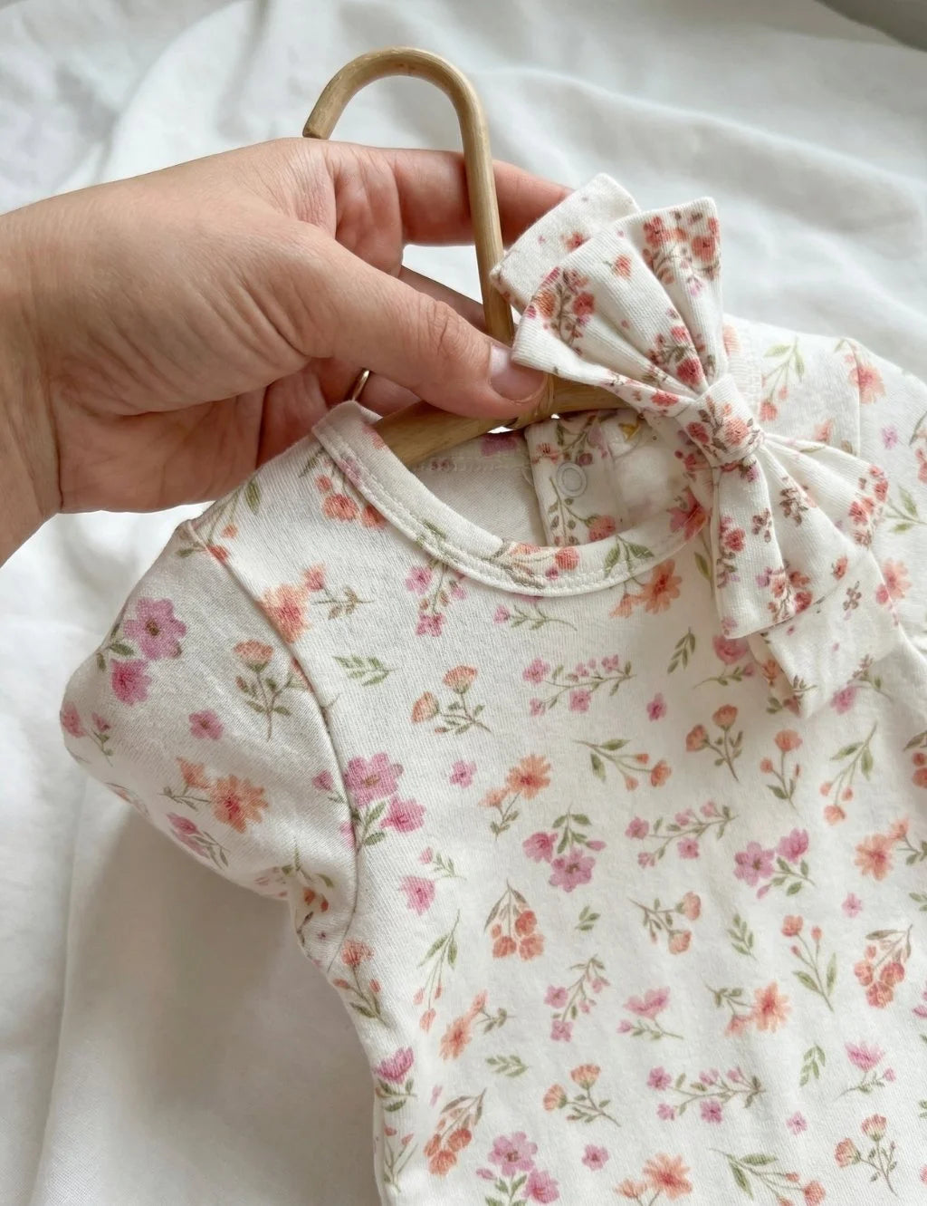 Close-up of a hand holding the pink floral organic cotton baby growsuit on a wooden hanger with the matching bow headband draped over the shoulder.