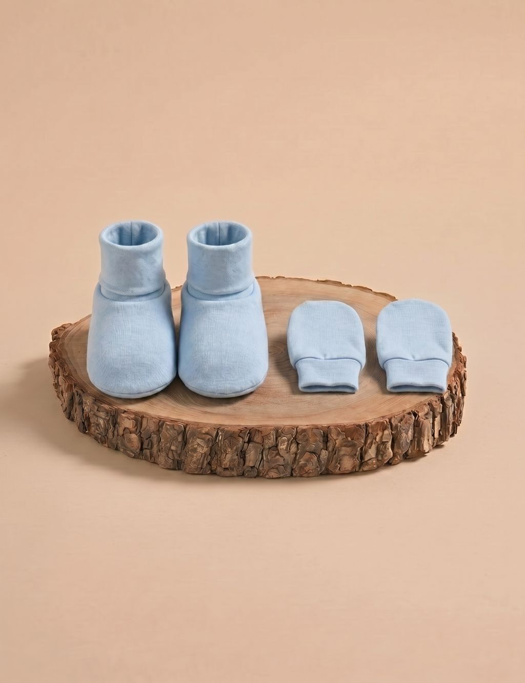 A matching set of solid light blue baby booties and scratch mittens, displayed on a rustic wooden log slice against a plain beige background.